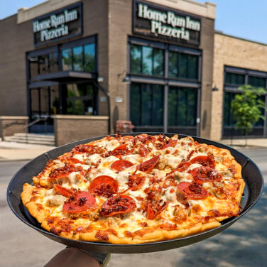 Pizza topped with sausage, bacon, and pepperoni outside held in front of restaurant