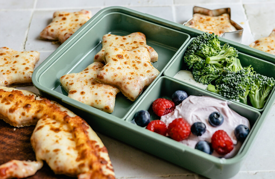 Pizza cut into star shapes and placed into a green lunchbox with mixed berries, yogurt, and broccoli on the side