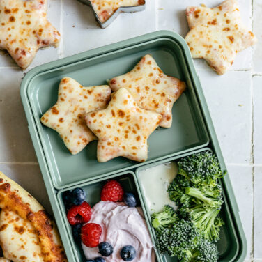 Pizza cut into star shapes and placed into a green lunchbox with mixed berries, yogurt, and broccoli on the side