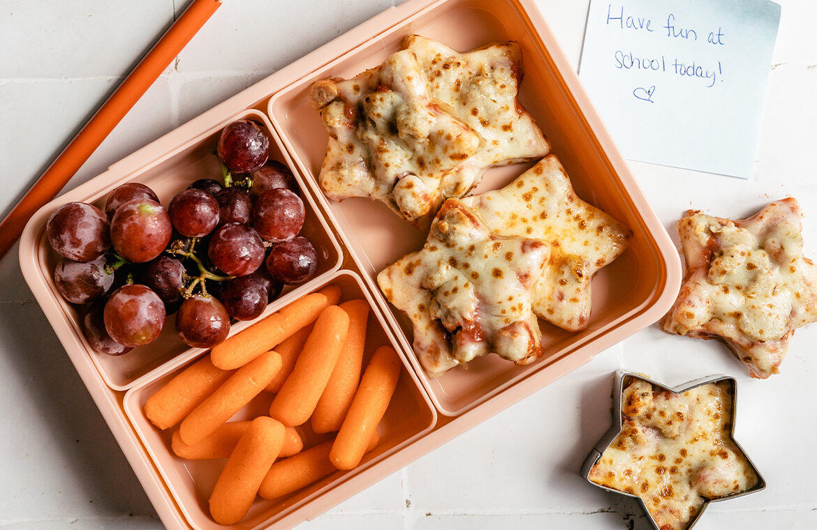 Pink lunchbox filled with sausage pizza cut into stars, baby carrots, red grapes, and marinara sauce on the side for dipping