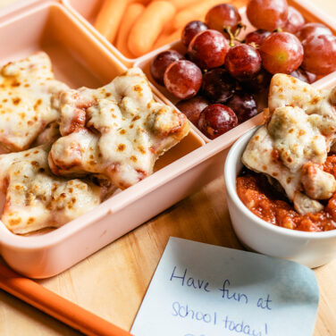 Pink lunchbox filled with sausage pizza cut into stars, baby carrots, red grapes, and marinara sauce on the side for dipping