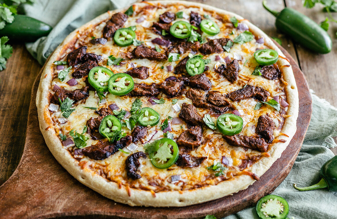 Pizza crust topped with Birria sauce, meat, jalapeños, and more