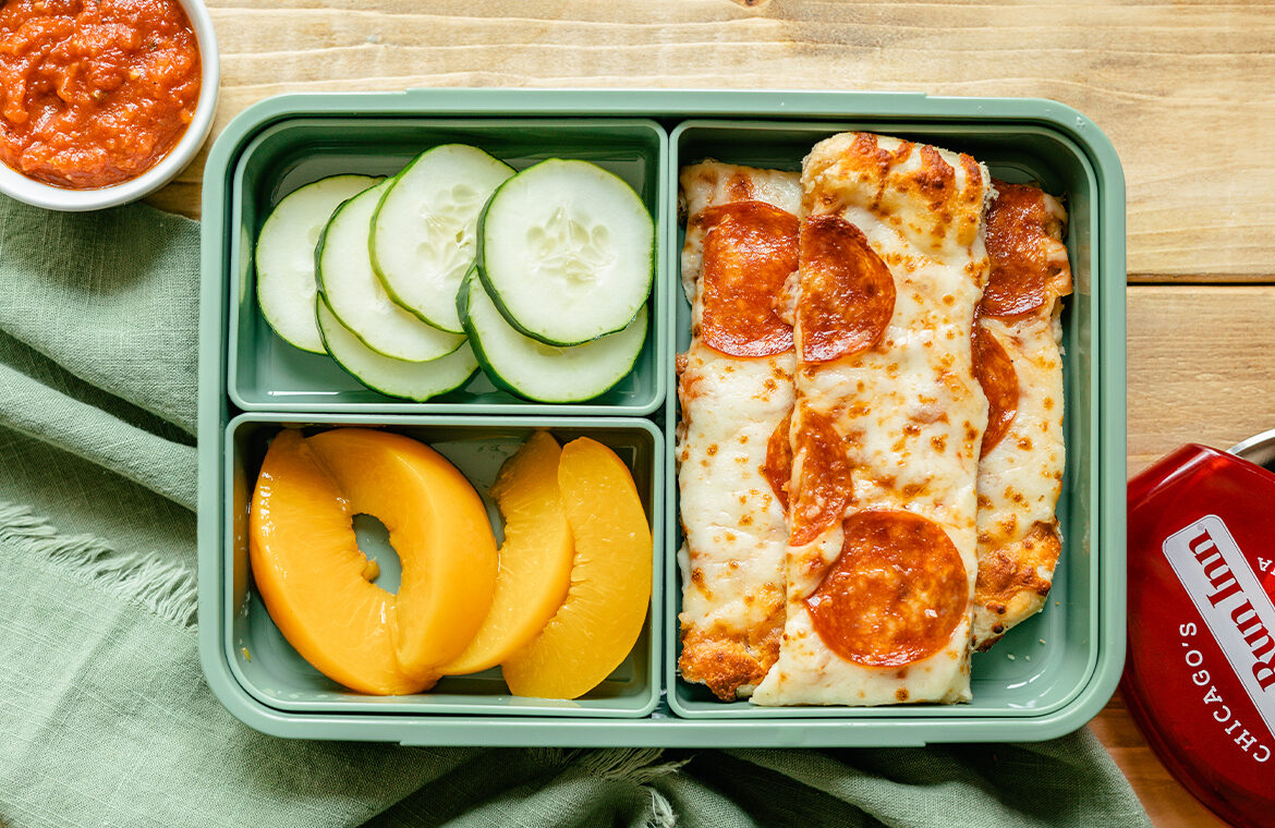 Green lunch box with pizza cut into strips, peaches, cucumbers, and marinara sauce for dipping