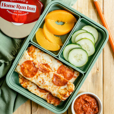 Green lunch box with pizza cut into strips, peaches, cucumbers, and marinara sauce for dipping