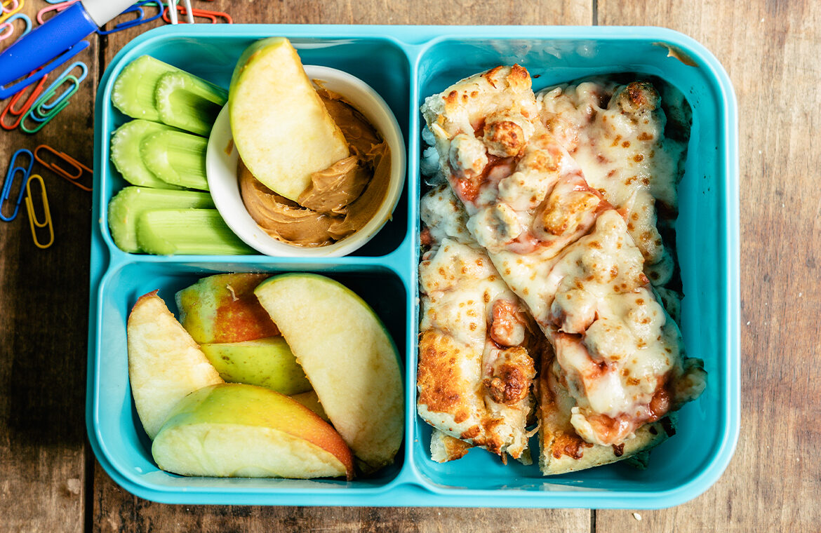 A blue llunchbox filled with sausage pizza cut into strips, celery sticks, sliced apples, and peanut butter