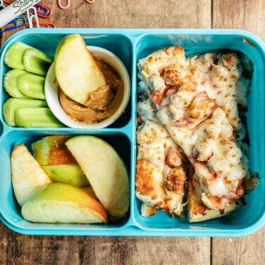A blue llunchbox filled with sausage pizza cut into strips, celery sticks, sliced apples, and peanut butter