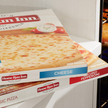 Two empty pizza boxes sitting on counter with cooked pizza coming out of the oven