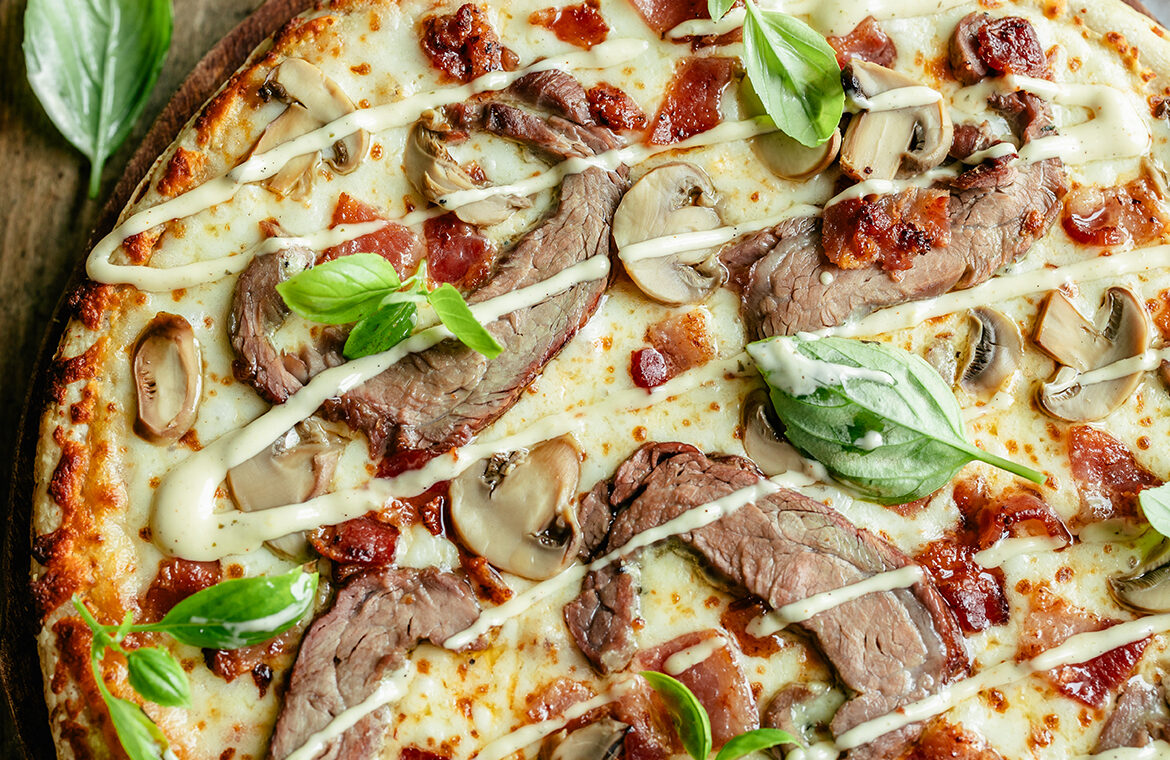 Cooked pizza topped with shaved steak, bacon, mushrooms, and garlic aioli drizzle