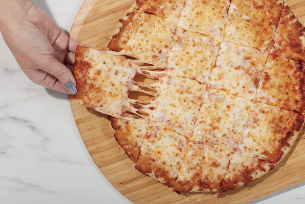 Hand pulling a square slice of Home Run Inn cheese thin crust pizza with melted cheese stretching on a wooden board.