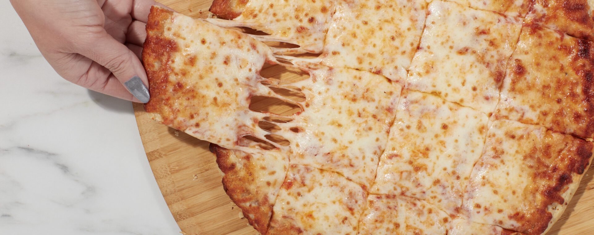 Hand pulling a square slice of Home Run Inn cheese thin crust pizza with melted cheese stretching on a wooden board.