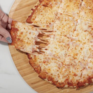 Hand pulling a square slice of Home Run Inn cheese thin crust pizza with melted cheese stretching on a wooden board.